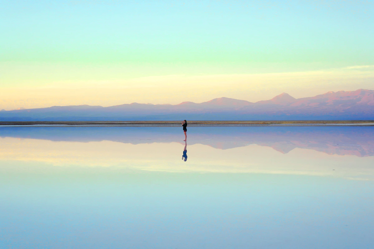 A person standing at the edge of a calm, mirror-still lake at dusk