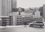 A monochrome image of an urban landscape featuring a multi-story car park adjacent to several tall office buildings. A lone minivan is parked in the foreground on what appears to be the rooftop of the parking structure.