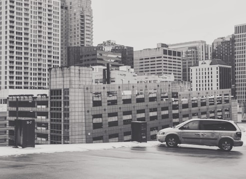 A monochrome image of an urban landscape featuring a multi-story car park adjacent to several tall office buildings. A lone minivan is parked in the foreground on what appears to be the rooftop of the parking structure.