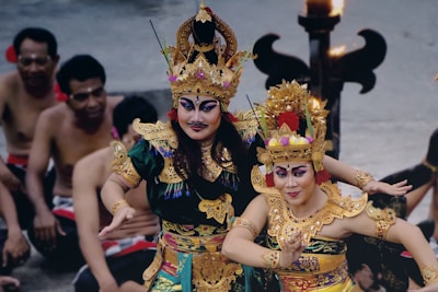 Two people are dressed in traditional Balinese dance costumes, adorned with intricate golden headpieces and colorful clothing. Their makeup is bold, emphasizing the eyes and facial expressions. They are positioned in a dance pose, with their arms elegantly extended. In the background, a group of people is seated, observing the performance, while a lit torch adds to the ambiance.