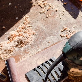 Wood shavings are scattered on a wooden surface, illuminated by sunlight. A power sander is positioned to the right, partially visible, suggesting a recent or ongoing sanding project. The scene is filled with the texture of sawdust and the contrasting smooth surface of sanded wood.