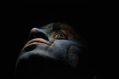 A close-up of a face with dramatic lighting, casting shadows that create a mysterious and intense atmosphere. The colors appear to be painted or enhanced, adding a surreal and artistic quality.