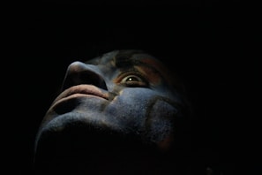 A close-up of a face with dramatic lighting, casting shadows that create a mysterious and intense atmosphere. The colors appear to be painted or enhanced, adding a surreal and artistic quality.
