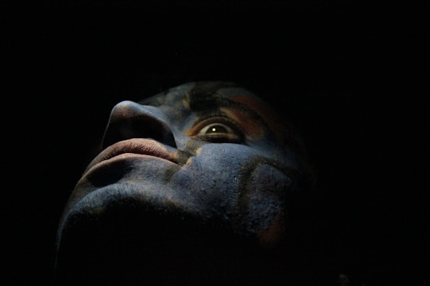 A close-up of a face with dramatic lighting, casting shadows that create a mysterious and intense atmosphere. The colors appear to be painted or enhanced, adding a surreal and artistic quality.