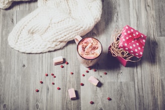 A cozy setting featuring a cream-colored knitted blanket draped over a wooden surface. Nearby, a mug filled with hot chocolate topped with marshmallows and sprinkled with chocolate powder sits alongside a small pink gift box adorned with white polka dots and a red bow. Scattered berries and marshmallows add to the warm and inviting atmosphere.