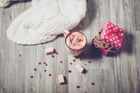 A cozy setting featuring a cream-colored knitted blanket draped over a wooden surface. Nearby, a mug filled with hot chocolate topped with marshmallows and sprinkled with chocolate powder sits alongside a small pink gift box adorned with white polka dots and a red bow. Scattered berries and marshmallows add to the warm and inviting atmosphere.