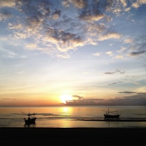 A serene seascape features a sunset over calm waters, with two small fishing boats silhouetted in the foreground. The sky is a mixture of golden, orange, and blue hues, scattered with soft clouds that reflect the sunset colors.