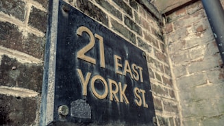 A weathered, metal street sign displaying the address '21 East York St.' is mounted on a brick wall. The surface of the sign shows signs of aging with rust and discoloration around the edges, and the wall has a rustic and textured appearance.