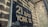 A weathered, metal street sign displaying the address '21 East York St.' is mounted on a brick wall. The surface of the sign shows signs of aging with rust and discoloration around the edges, and the wall has a rustic and textured appearance.