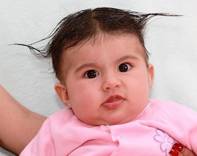 A baby with dark eyes and wet hair styled to stand up on the sides is wearing a pink outfit with a flower design. The infant is looking directly at the camera while lying on a light-colored surface.