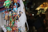 Colorful fabric ornaments shaped like animals are hanging closely together, adorned with beads and small bells. The background is blurred, suggesting a market setting with various other crafts and merchandise.