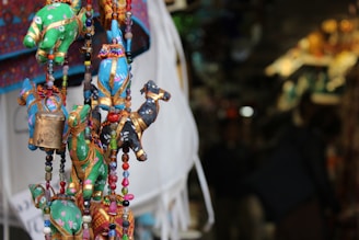 Colorful fabric ornaments shaped like animals are hanging closely together, adorned with beads and small bells. The background is blurred, suggesting a market setting with various other crafts and merchandise.