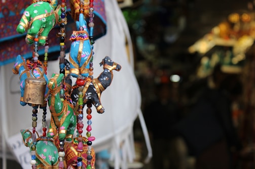 Colorful fabric ornaments shaped like animals are hanging closely together, adorned with beads and small bells. The background is blurred, suggesting a market setting with various other crafts and merchandise.