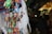 Colorful fabric ornaments shaped like animals are hanging closely together, adorned with beads and small bells. The background is blurred, suggesting a market setting with various other crafts and merchandise.