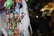 Colorful fabric ornaments shaped like animals are hanging closely together, adorned with beads and small bells. The background is blurred, suggesting a market setting with various other crafts and merchandise.
