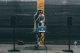 A DJ stands behind a small table setup with a laptop and audio equipment. Large black speakers on stands are positioned on either side. The DJ wears headphones and holds a microphone, performing in front of a backdrop of dark metal shutters with a single streak of yellow paint in the center.