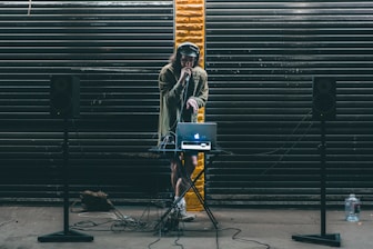 A DJ stands behind a small table setup with a laptop and audio equipment. Large black speakers on stands are positioned on either side. The DJ wears headphones and holds a microphone, performing in front of a backdrop of dark metal shutters with a single streak of yellow paint in the center.
