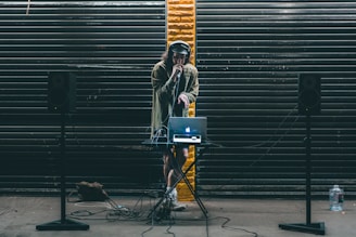 A DJ stands behind a small table setup with a laptop and audio equipment. Large black speakers on stands are positioned on either side. The DJ wears headphones and holds a microphone, performing in front of a backdrop of dark metal shutters with a single streak of yellow paint in the center.