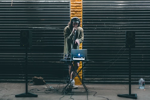 A DJ stands behind a small table setup with a laptop and audio equipment. Large black speakers on stands are positioned on either side. The DJ wears headphones and holds a microphone, performing in front of a backdrop of dark metal shutters with a single streak of yellow paint in the center.