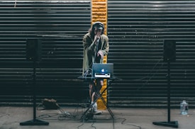 A DJ stands behind a small table setup with a laptop and audio equipment. Large black speakers on stands are positioned on either side. The DJ wears headphones and holds a microphone, performing in front of a backdrop of dark metal shutters with a single streak of yellow paint in the center.