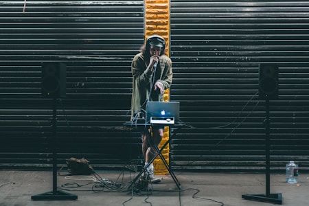 A DJ stands behind a small table setup with a laptop and audio equipment. Large black speakers on stands are positioned on either side. The DJ wears headphones and holds a microphone, performing in front of a backdrop of dark metal shutters with a single streak of yellow paint in the center.