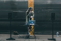 A DJ stands behind a small table setup with a laptop and audio equipment. Large black speakers on stands are positioned on either side. The DJ wears headphones and holds a microphone, performing in front of a backdrop of dark metal shutters with a single streak of yellow paint in the center.