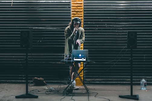 A DJ stands behind a small table setup with a laptop and audio equipment. Large black speakers on stands are positioned on either side. The DJ wears headphones and holds a microphone, performing in front of a backdrop of dark metal shutters with a single streak of yellow paint in the center.