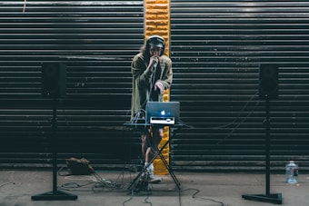 A DJ stands behind a small table setup with a laptop and audio equipment. Large black speakers on stands are positioned on either side. The DJ wears headphones and holds a microphone, performing in front of a backdrop of dark metal shutters with a single streak of yellow paint in the center.