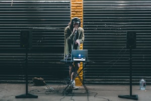 A DJ stands behind a small table setup with a laptop and audio equipment. Large black speakers on stands are positioned on either side. The DJ wears headphones and holds a microphone, performing in front of a backdrop of dark metal shutters with a single streak of yellow paint in the center.