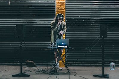A DJ stands behind a small table setup with a laptop and audio equipment. Large black speakers on stands are positioned on either side. The DJ wears headphones and holds a microphone, performing in front of a backdrop of dark metal shutters with a single streak of yellow paint in the center.
