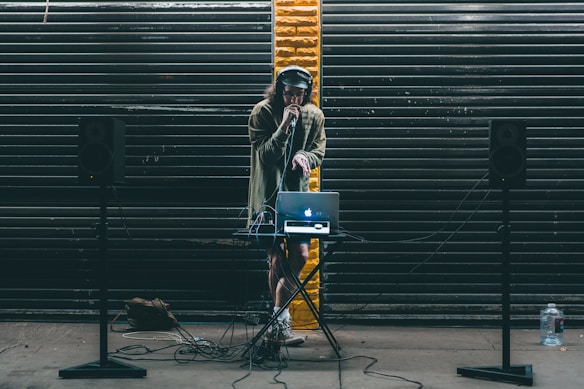 A DJ stands behind a small table setup with a laptop and audio equipment. Large black speakers on stands are positioned on either side. The DJ wears headphones and holds a microphone, performing in front of a backdrop of dark metal shutters with a single streak of yellow paint in the center.