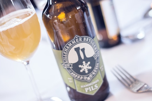 A glass of pale beer sits next to a bottle labeled 'Lillehammer Bryggeri Hammonds Pils' on a white tablecloth. The scene includes a metallic fork nearby, and another glass bottle blurred in the background.
