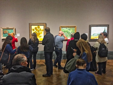 A group of people are gathered in an art gallery, observing several framed paintings on a wall. The paintings vary in style and include a depiction of sunflowers and serene landscapes. People are standing and some are using their phones, possibly taking pictures or notes. The room has a quiet and contemplative atmosphere, with a wooden floor and neutral-colored walls.