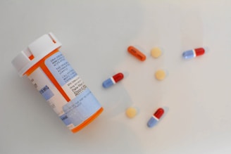 A plastic prescription bottle with a white cap and blue label lies on its side alongside assorted pills. The pills include small round yellow tablets, red and blue capsules, and an orange and white capsule.