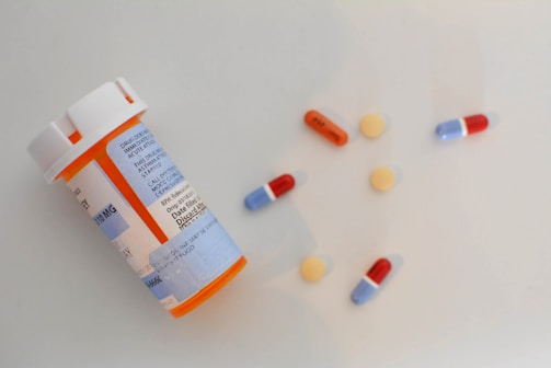 A plastic prescription bottle with a white cap and blue label lies on its side alongside assorted pills. The pills include small round yellow tablets, red and blue capsules, and an orange and white capsule.