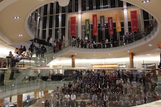 A bustling indoor event featuring a large group of people gathered in a modern, multi-level atrium with glass railings. The crowd is distributed across different levels of the venue, some sitting on steps and others standing. Colorful banners with words like Integrity, Collaboration, and Excellence hang from the upper floor.