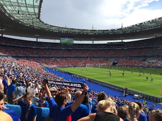 A large stadium filled with spectators wearing blue and red clothing, with a soccer match taking place on the field. The atmosphere appears lively and energetic, with fans joyfully waving scarves and cheering. Bright sunlight illuminates the scene, casting shadows across the seating areas.