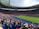 A large stadium filled with spectators wearing blue and red clothing, with a soccer match taking place on the field. The atmosphere appears lively and energetic, with fans joyfully waving scarves and cheering. Bright sunlight illuminates the scene, casting shadows across the seating areas.