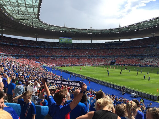A large stadium filled with spectators wearing blue and red clothing, with a soccer match taking place on the field. The atmosphere appears lively and energetic, with fans joyfully waving scarves and cheering. Bright sunlight illuminates the scene, casting shadows across the seating areas.