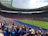 A large stadium filled with spectators wearing blue and red clothing, with a soccer match taking place on the field. The atmosphere appears lively and energetic, with fans joyfully waving scarves and cheering. Bright sunlight illuminates the scene, casting shadows across the seating areas.