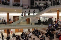 A large indoor space with multiple levels connected by glass railings. Crowds of people are dispersed throughout, some seated on stepped seating areas, others standing or walking. It’s a busy, social atmosphere with groups engaged in conversation. Pillars and modern architectural elements are visible.