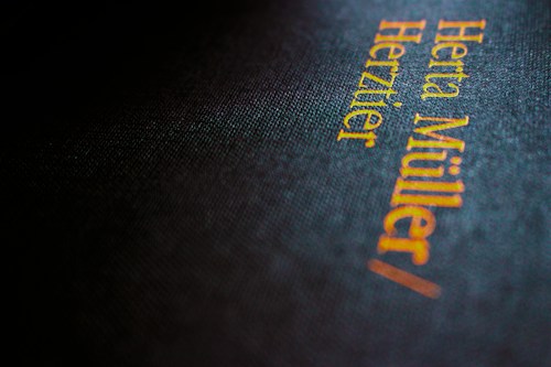 Close-up view of a book cover with textured black material, featuring gold and orange embossed text reading 'Herta M&uuml;ller' and 'Herztier.' The image is slightly out of focus, giving it a soft, moody feel.
