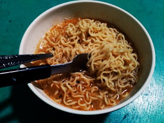 A bowl of instant noodles with broth, accompanied by a fork and a pair of chopsticks resting on the bowl's edge. The background is a textured teal surface.