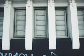 A row of rectangular windows is set into a wall with white vertical beams dividing each window section. The lower portion of the wall is painted dark, with some graffiti visible at the bottom.