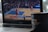 A laptop displays a blurred basketball game in progress on its screen. In the foreground, a black cup with a logo and a wooden stirrer sits on the edge of the laptop.