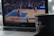 A laptop displays a blurred basketball game in progress on its screen. In the foreground, a black cup with a logo and a wooden stirrer sits on the edge of the laptop.