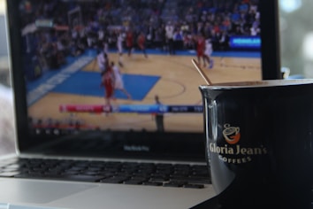 A laptop displays a blurred basketball game in progress on its screen. In the foreground, a black cup with a logo and a wooden stirrer sits on the edge of the laptop.
