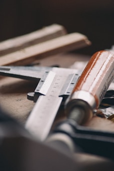 A close-up of various woodworking tools on a workbench, including a ruler and a chisel with a wooden handle. The tools have a slightly worn look, indicating regular use, and are arranged in a casual manner.