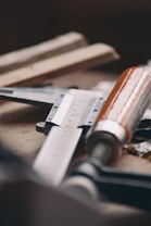 A close-up of various woodworking tools on a workbench, including a ruler and a chisel with a wooden handle. The tools have a slightly worn look, indicating regular use, and are arranged in a casual manner.