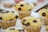 A close-up view of several freshly baked muffins with chocolate chips on top. The muffins are placed in white paper liners and are arranged on a light-colored surface. In the blurred background, there are additional baked goods, possibly macarons.